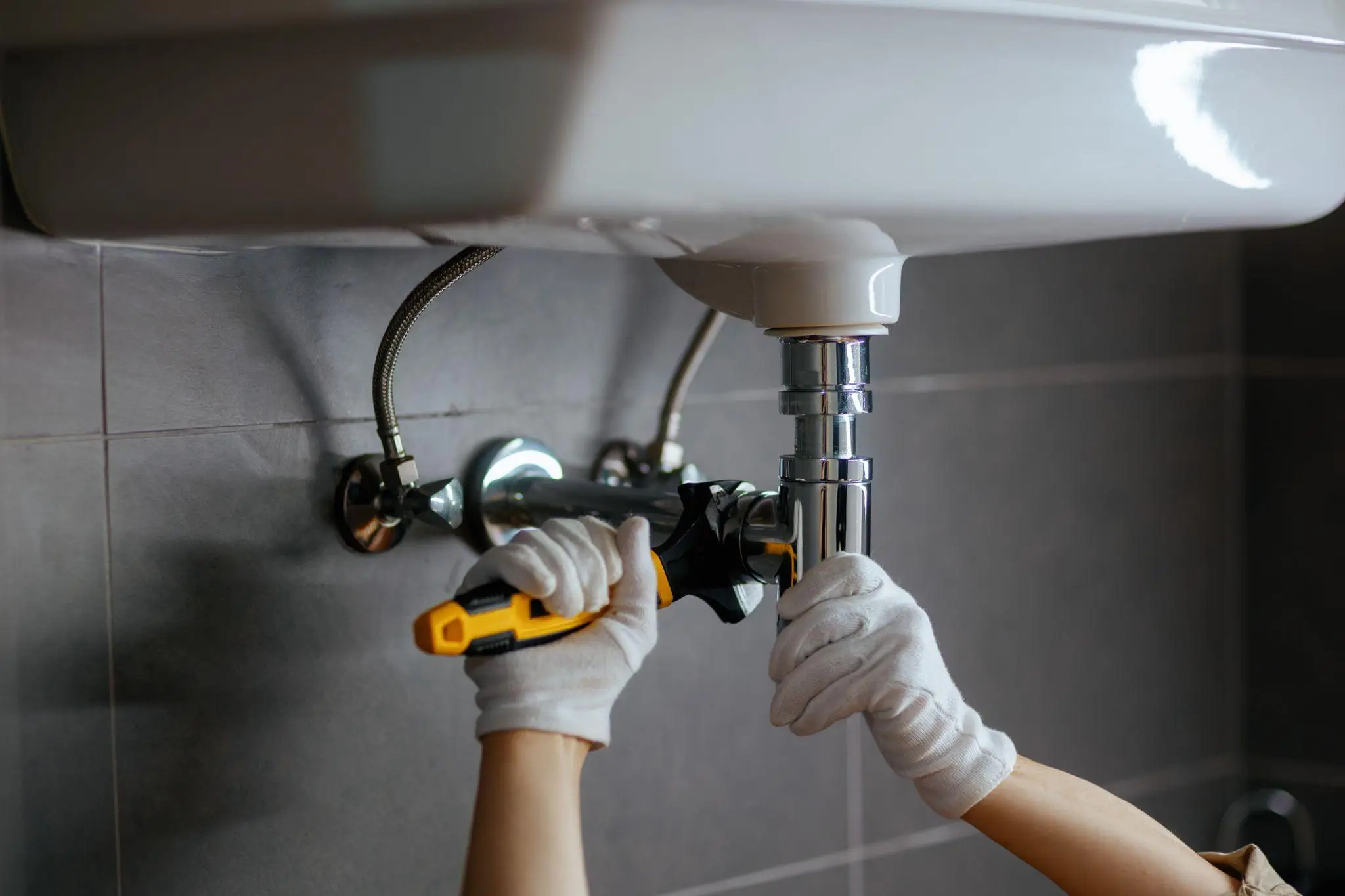 Gloved hands using wrench to tighten pipe under bathroom sink with gray tiled wall