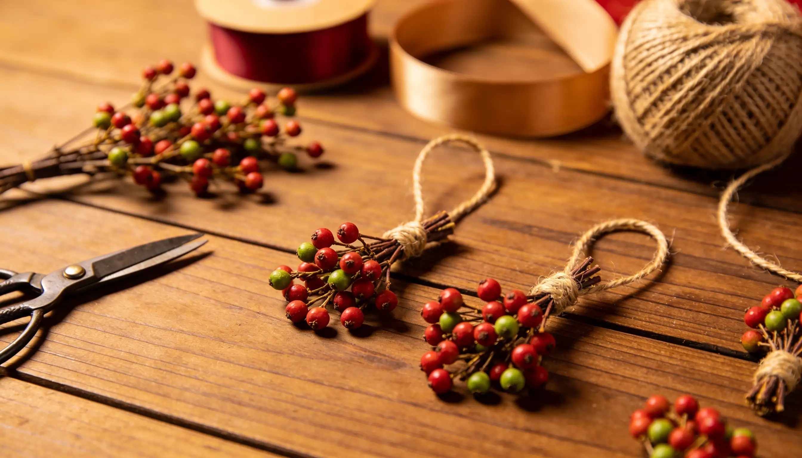 Craft supplies including berry sprigs, scissors, twine, and ribbons are arranged on a wooden surface.