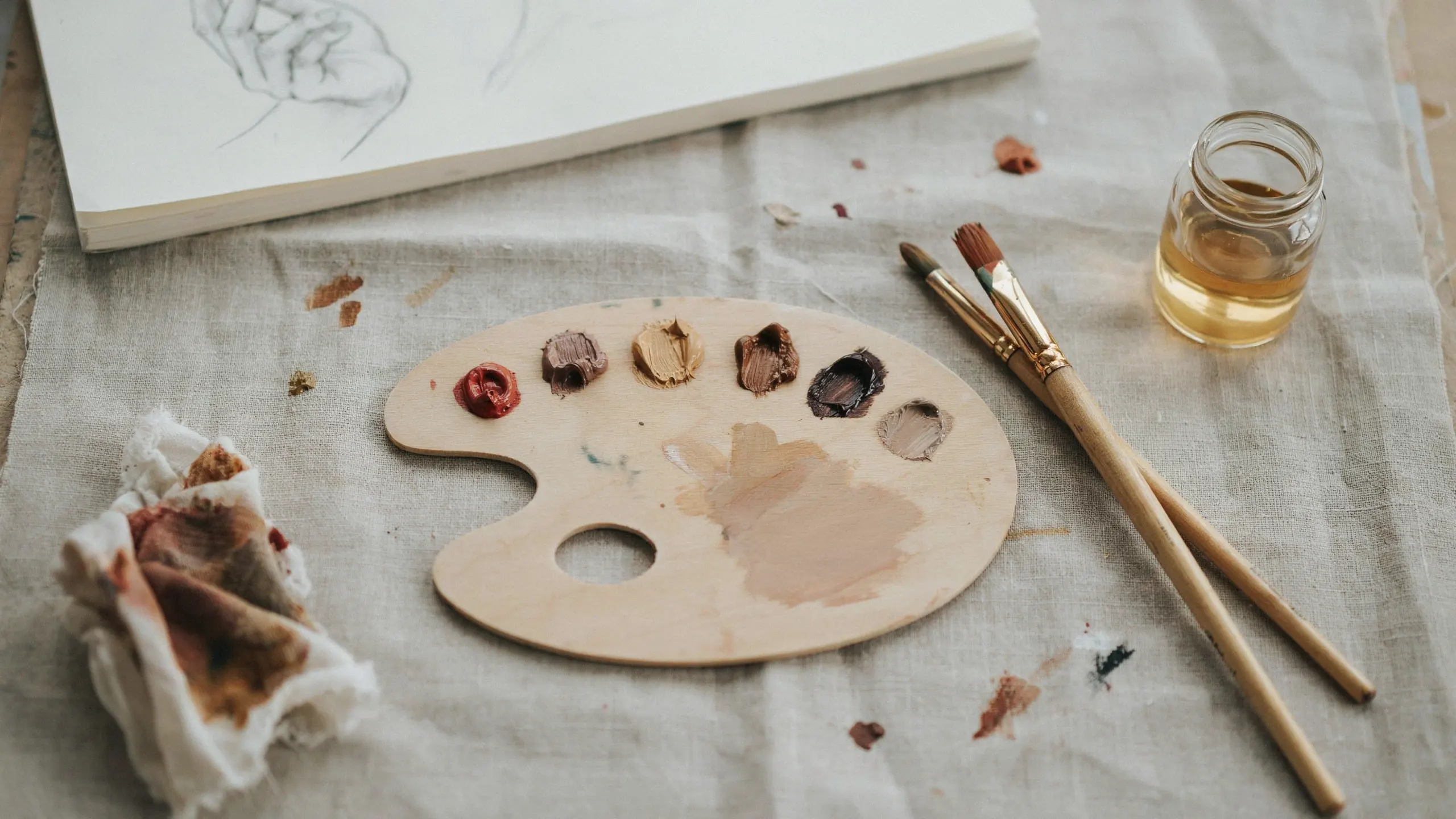 A wooden table displays a paint palette, brushes, and an open book, suggesting an artistic workspace.