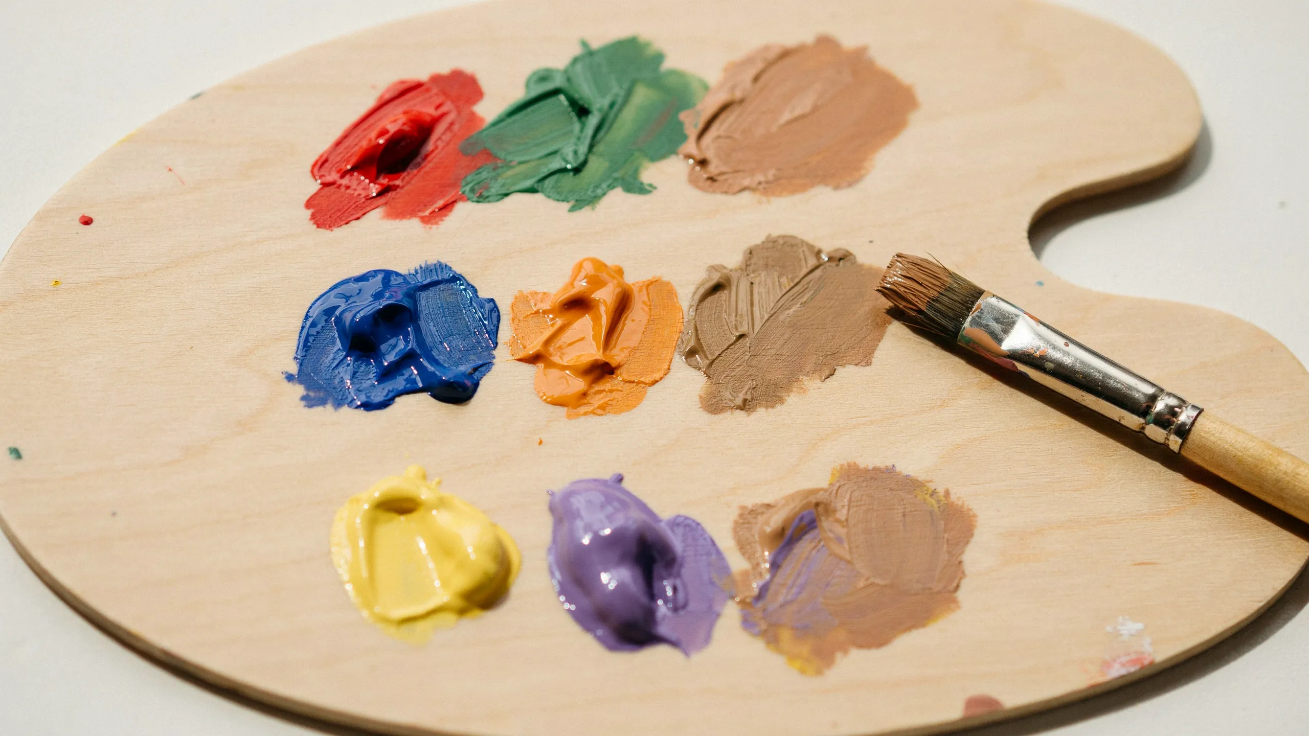 Wooden palette with colorful complementary paint blobs and a brush beside showing mixed shades of brown paint.