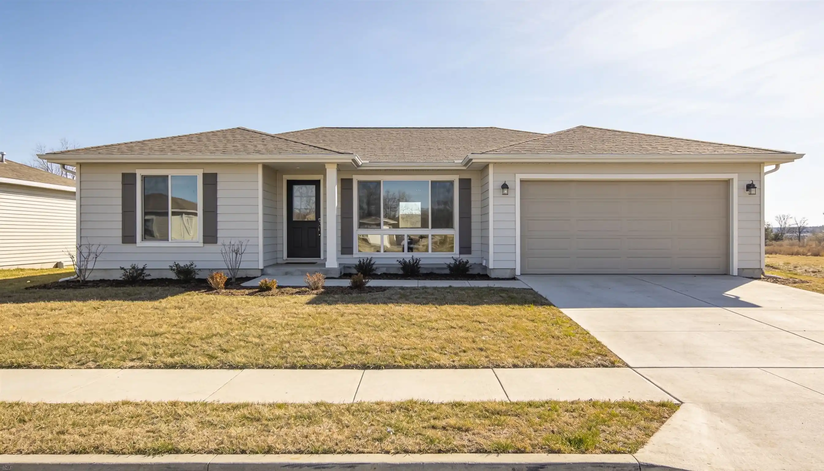 A light gray, single-story house with a dark front door, large front windows, and an attached garage sits under a clear blue sky.