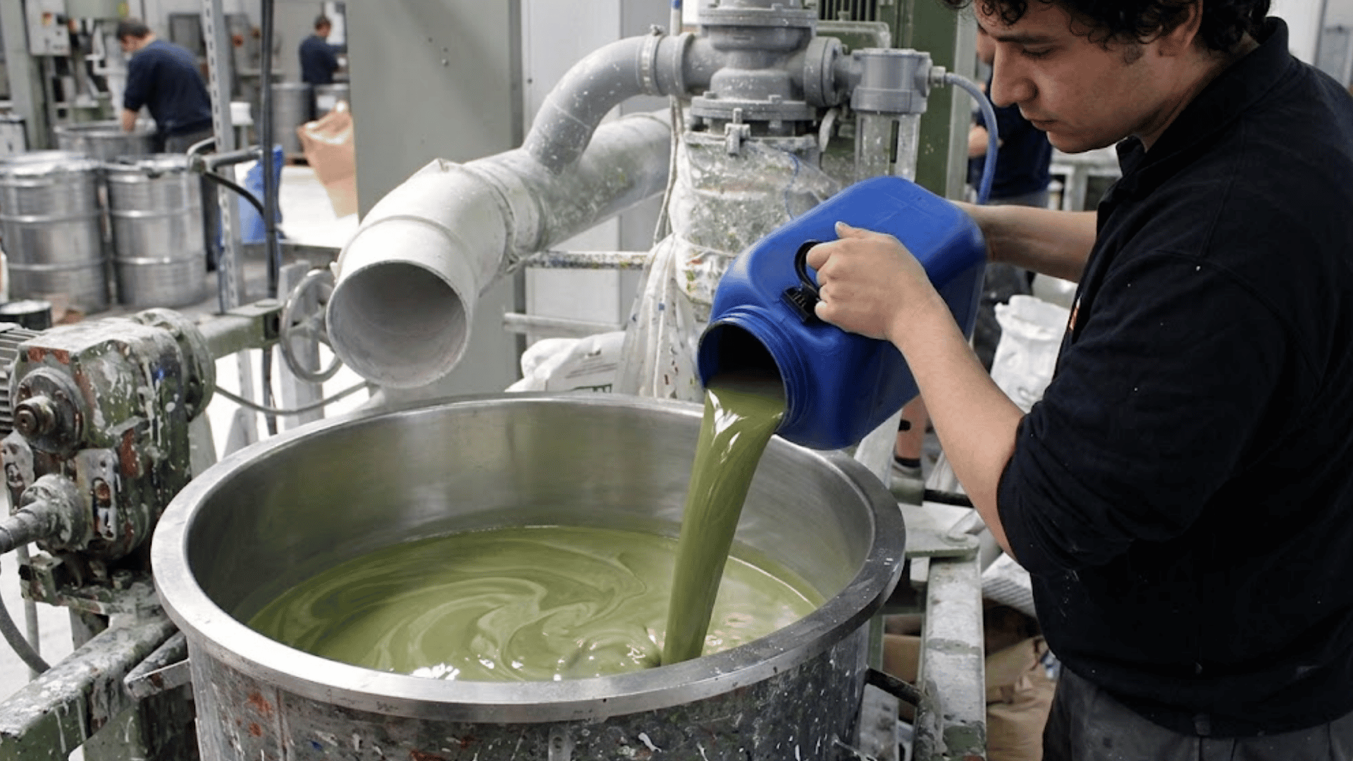 factory worker pours green paint into a large mixing vat during the industrial paint production process