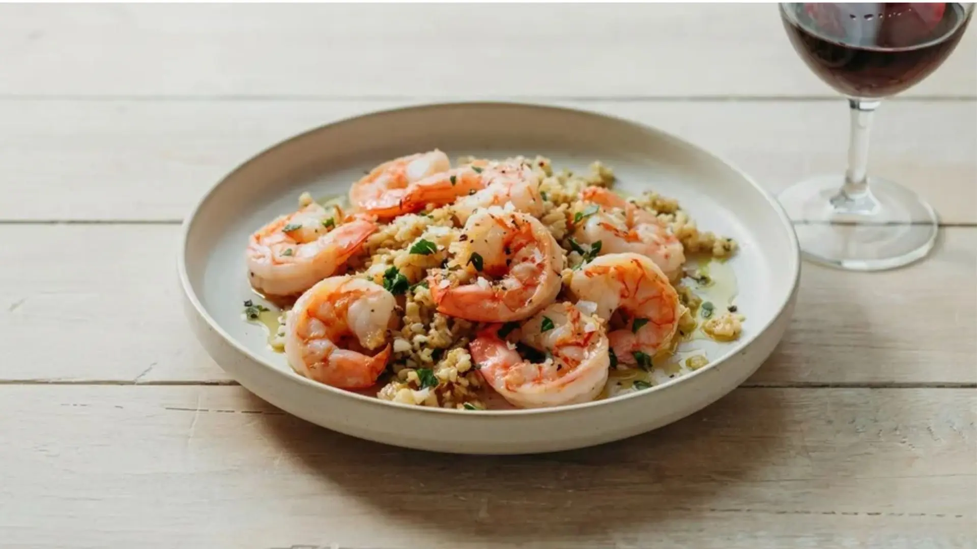 Plate of cooked shrimp with chopped herbs served over a bed of grains with a glass of red wine.