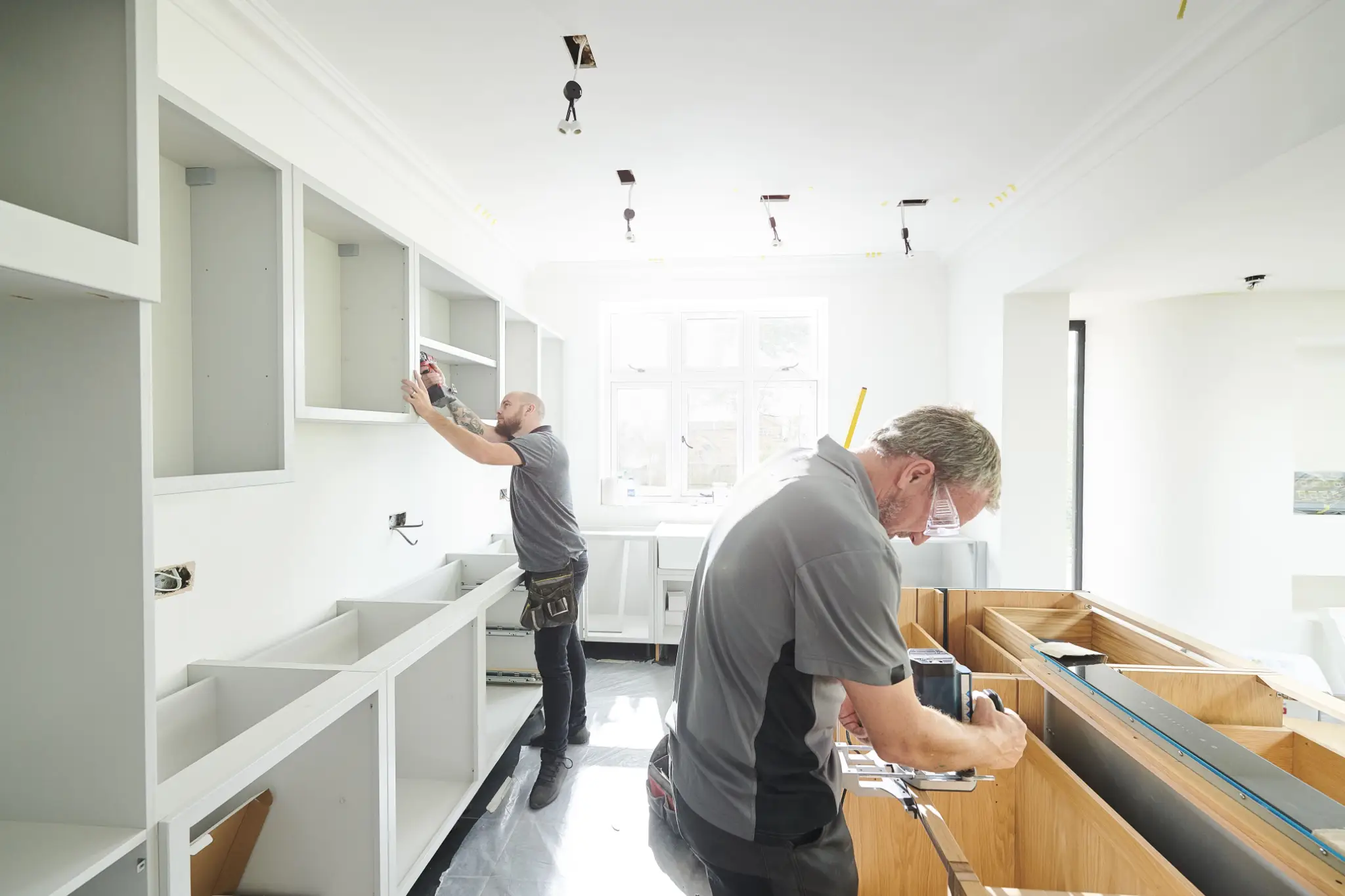 Two workers installing kitchen cabinets in a bright, unfinished room with power tools