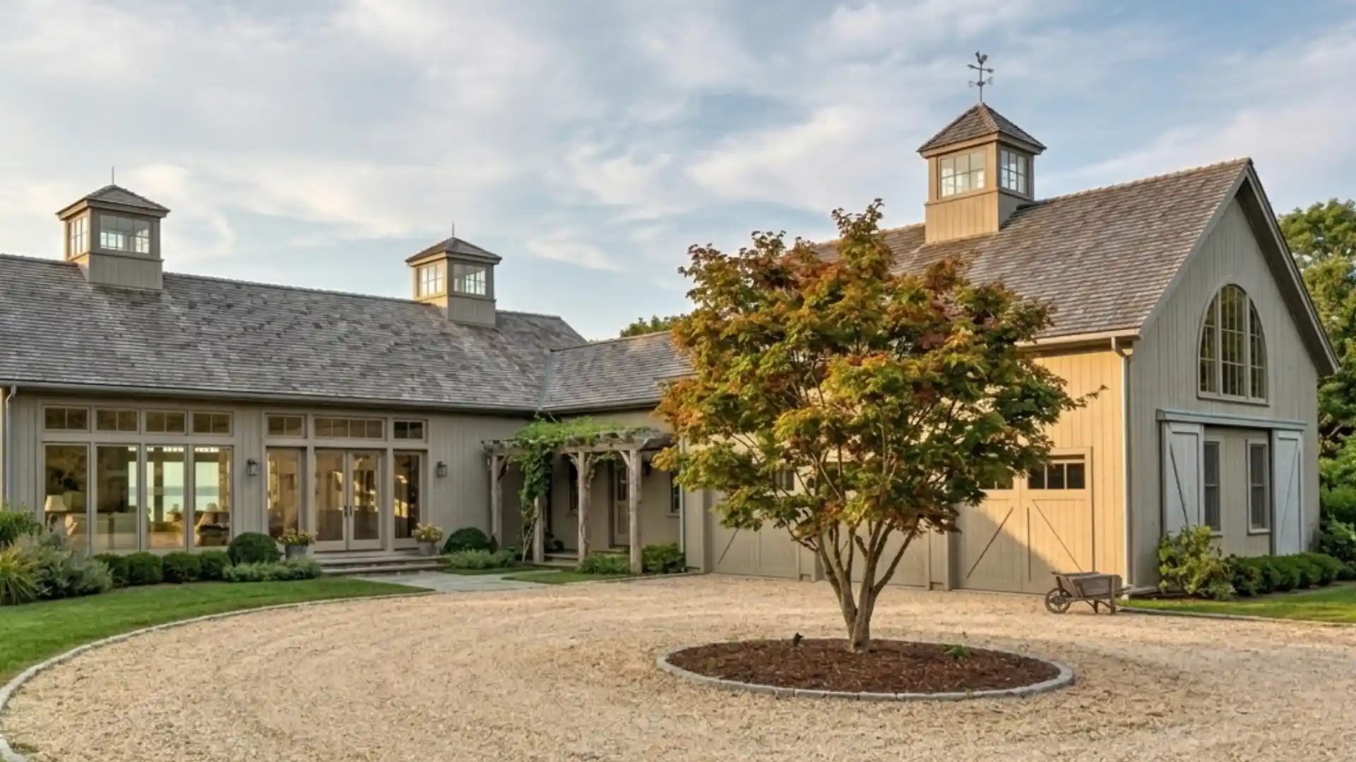An L shaped rambler house with dormer windows and a weather vane sits on a gravel driveway with a small tree in the foreground.