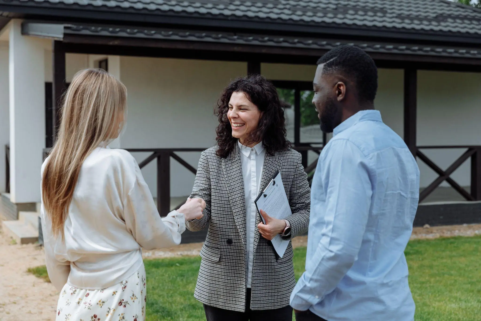Three people meeting outside a house, holding documents and shaking hands on a sunny day