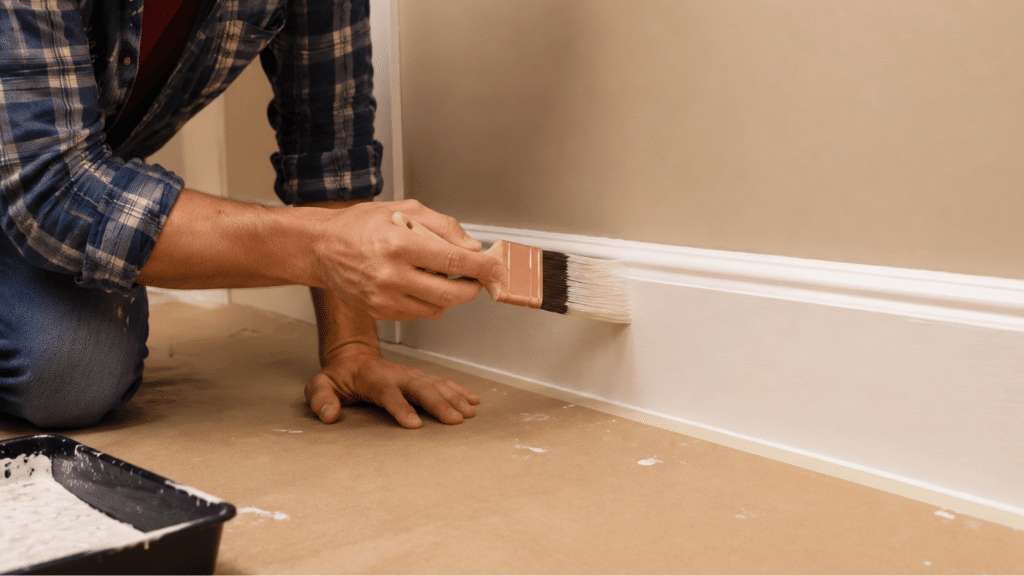 person painting white baseboard trim with brush during a home wall painting project indoors