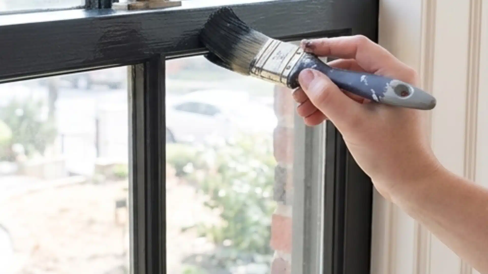 Hand painting a dark window frame with a brush.