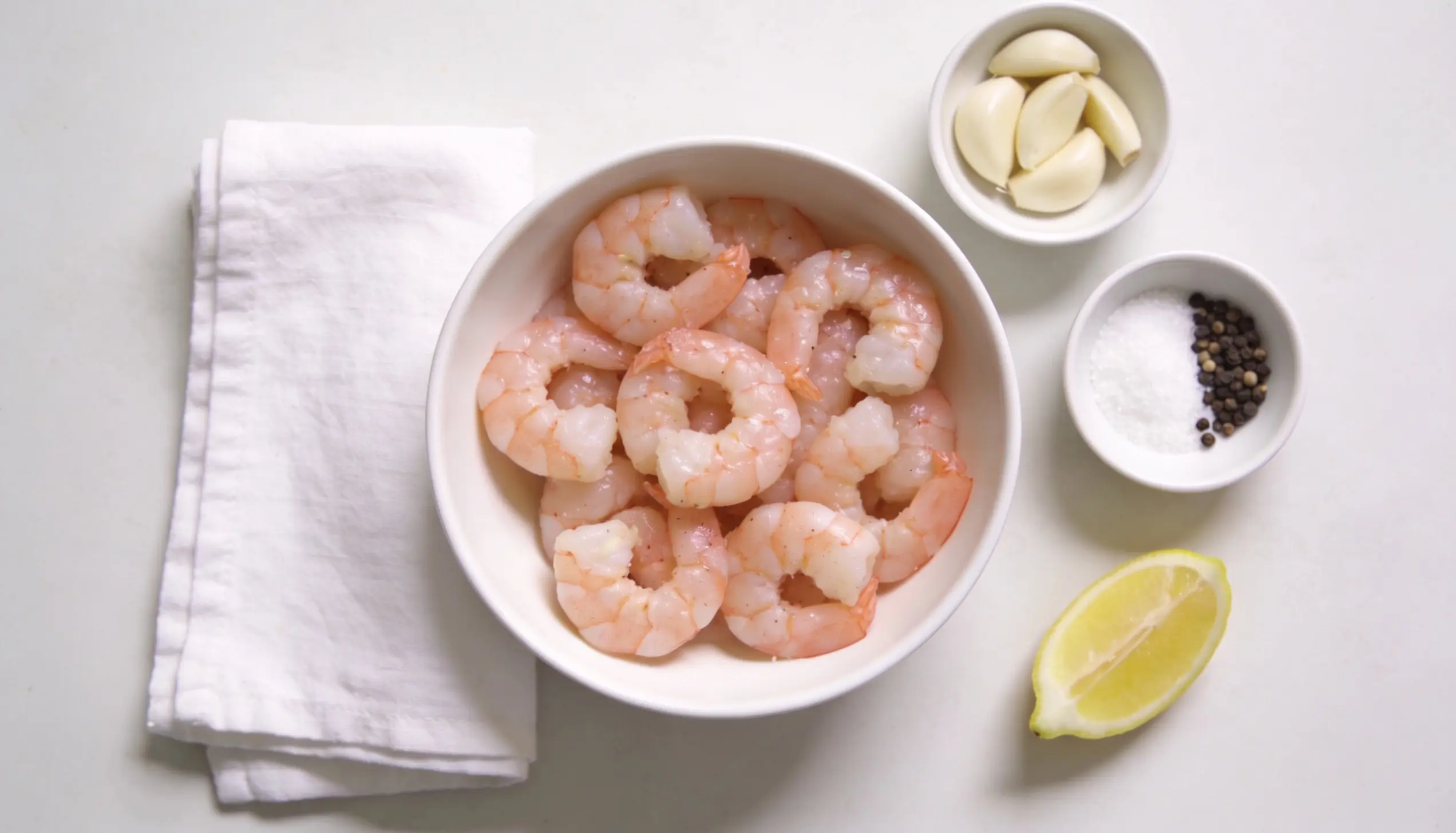 Bowl of shrimp with garlic cloves, salt, pepper, and a lemon wedge.