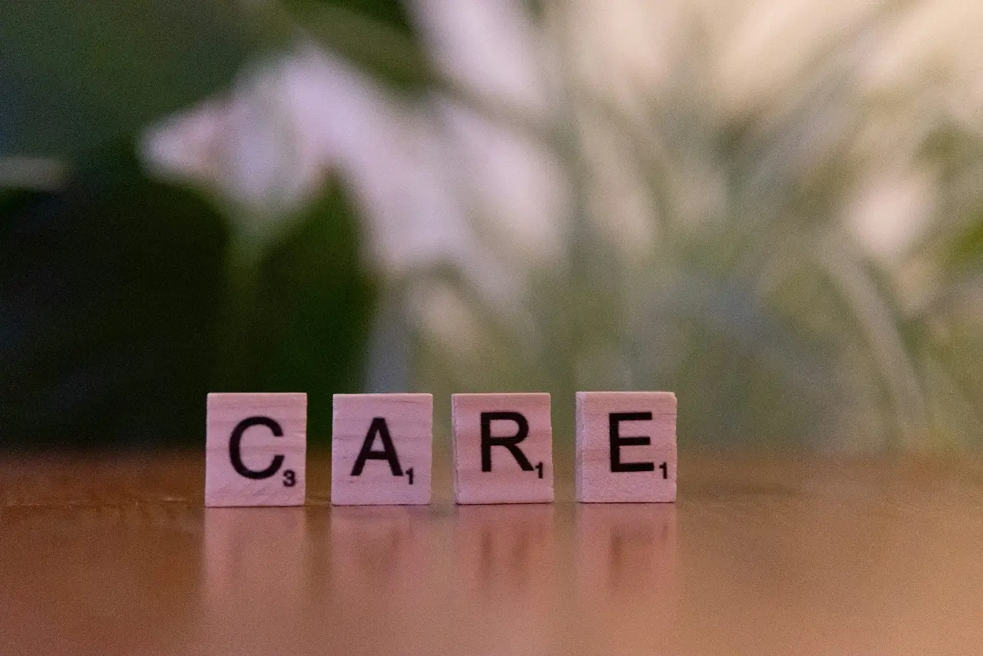Scrabble tiles spelling "CARE" on wooden surface with blurred plants in background