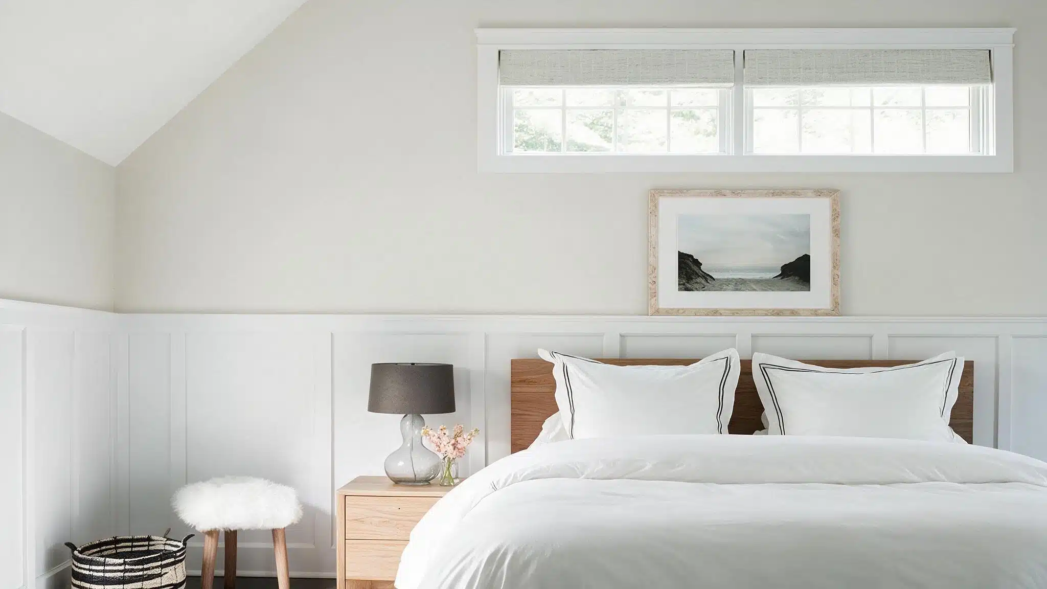 A bright bedroom with a neatly made bed, wood nightstand, lamp, flowers, stool, basket, framed landscape, and high windows.