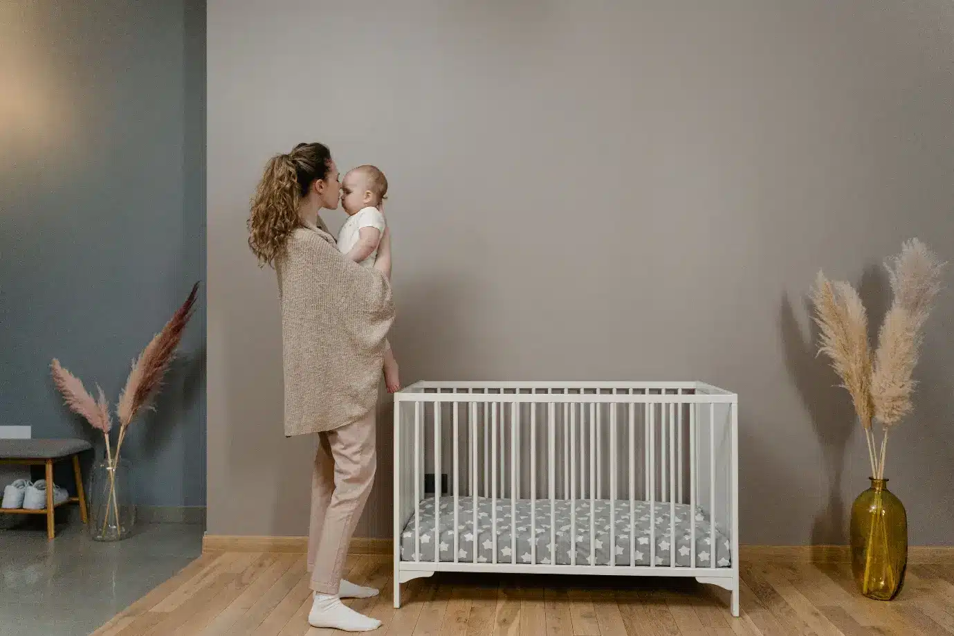 Mother holding baby next to white crib in cozy nursery with pampas grass decor