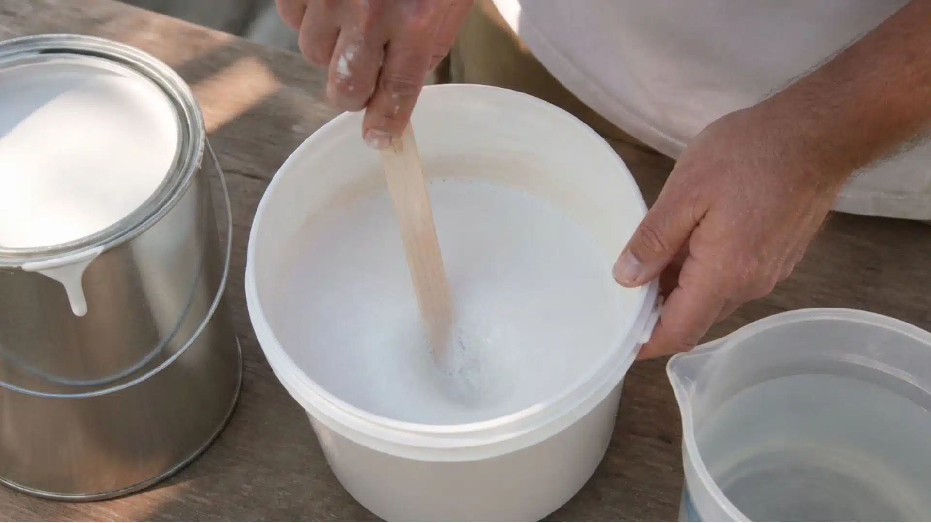 A person stirs a white substance in a bucket using a large mixing tool.