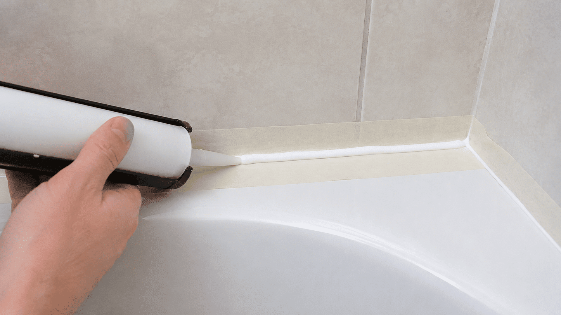 hand using a caulk gun to apply a fresh white bead along a taped bathtub and tiled wall joint