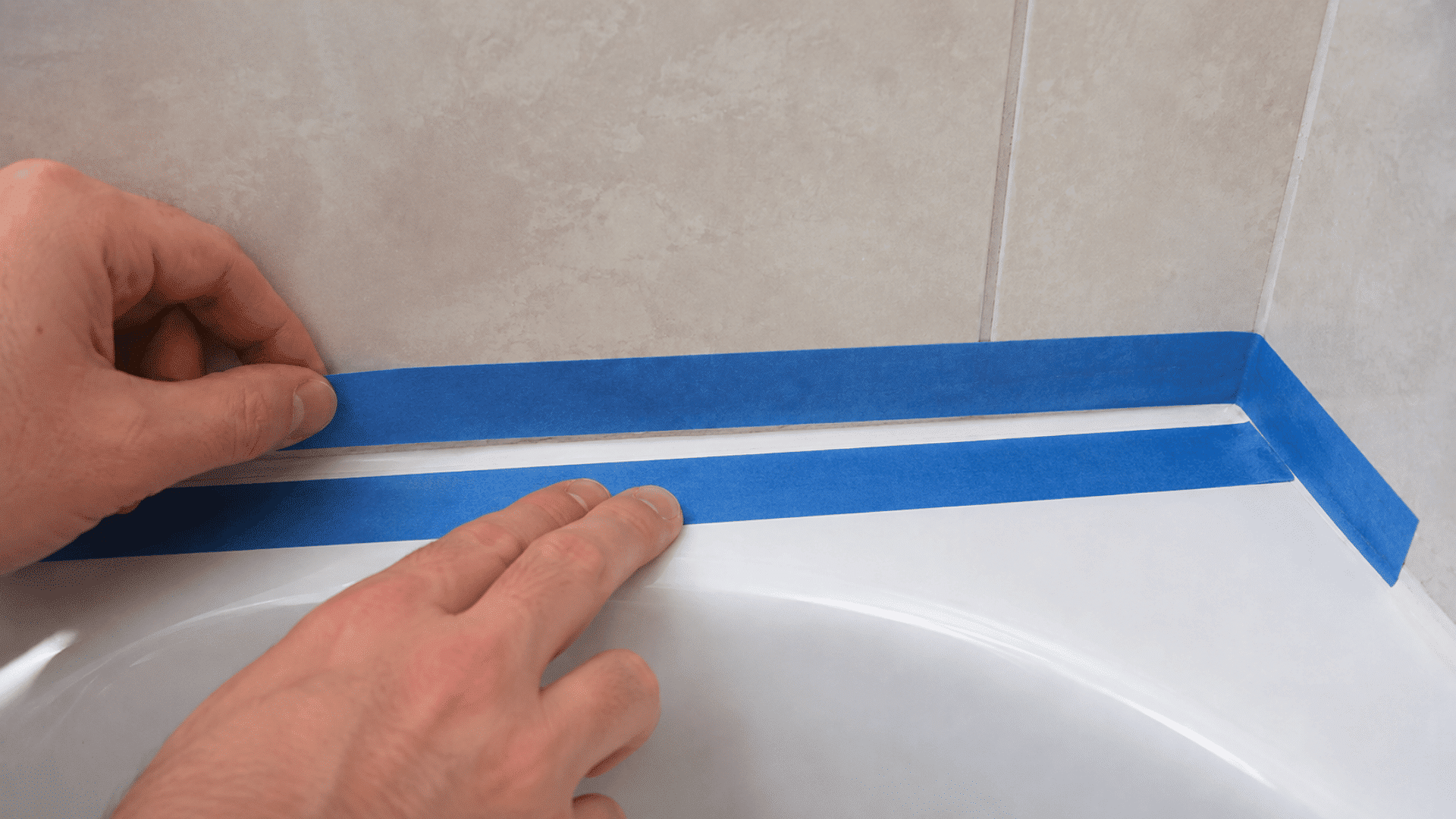 hands applying blue painter's tape above and below the joint between a white bathtub and tiled wall