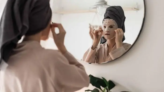 Woman applying facial sheet mask in front of circular bathroom mirror with towel on head