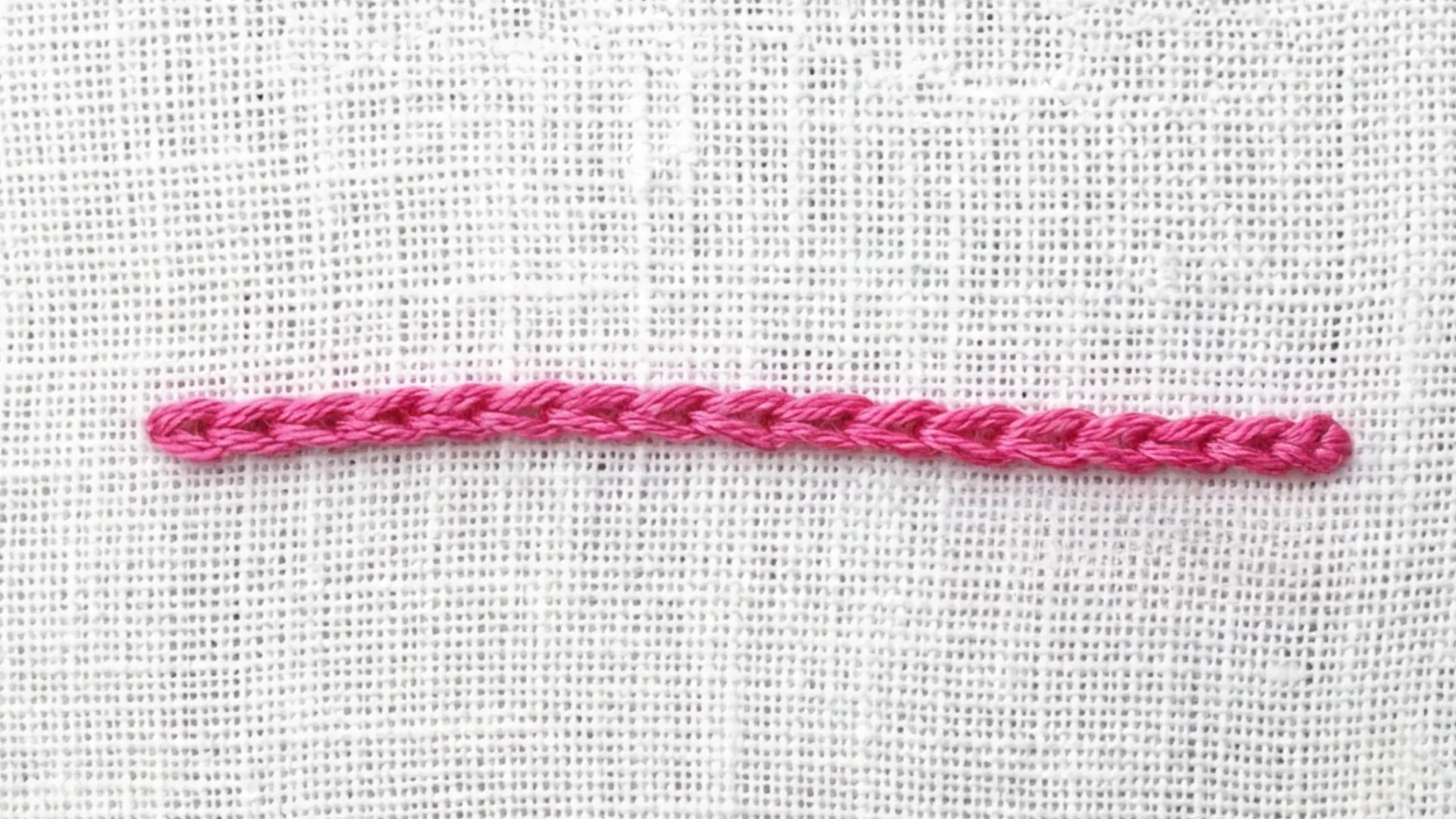 Chain stitch in pink thread forming a straight line of closely linked loops across textured white embroidery fabric