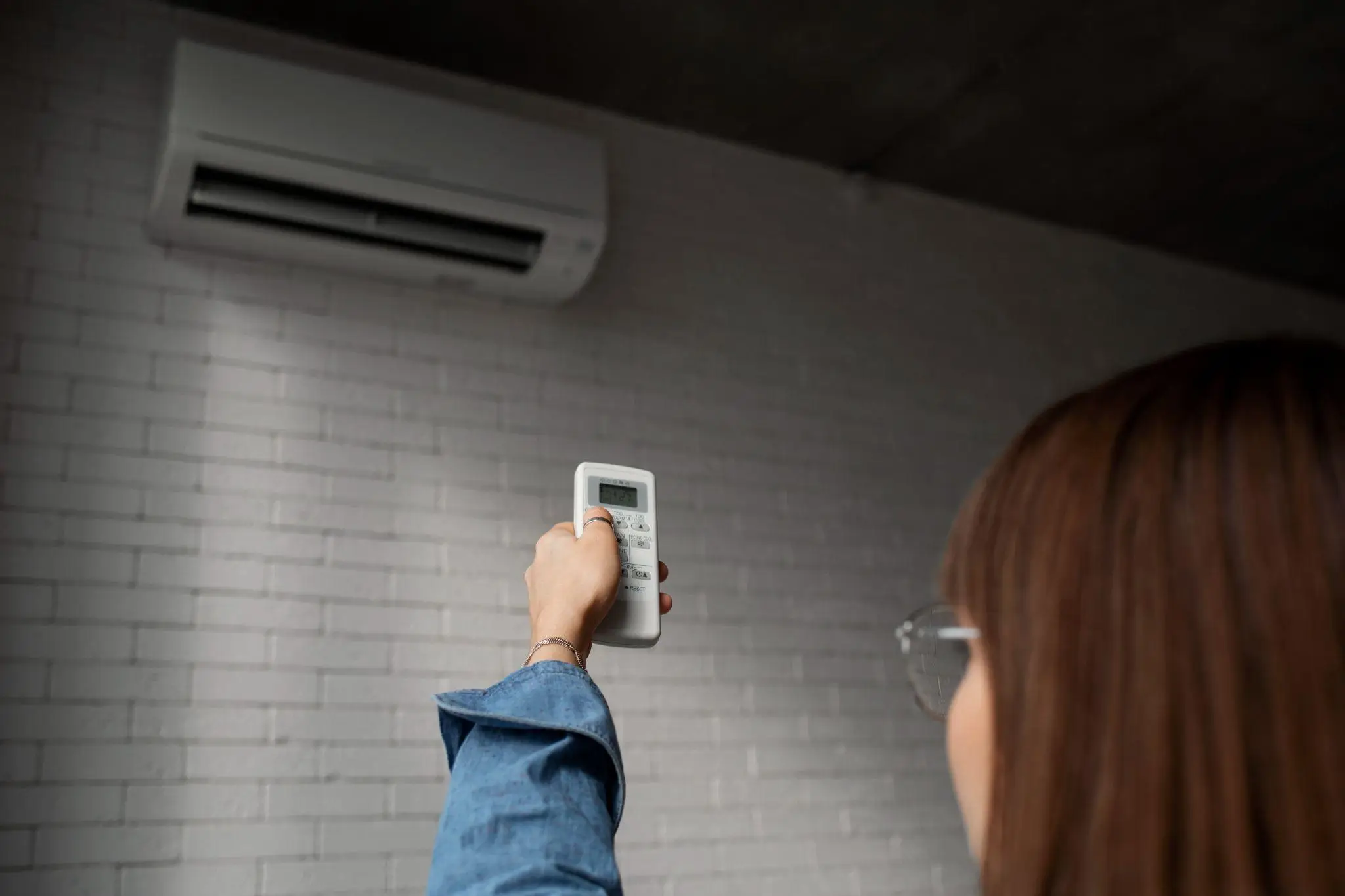 Person adjusting wall-mounted air conditioner with remote in a room with white brick walls
