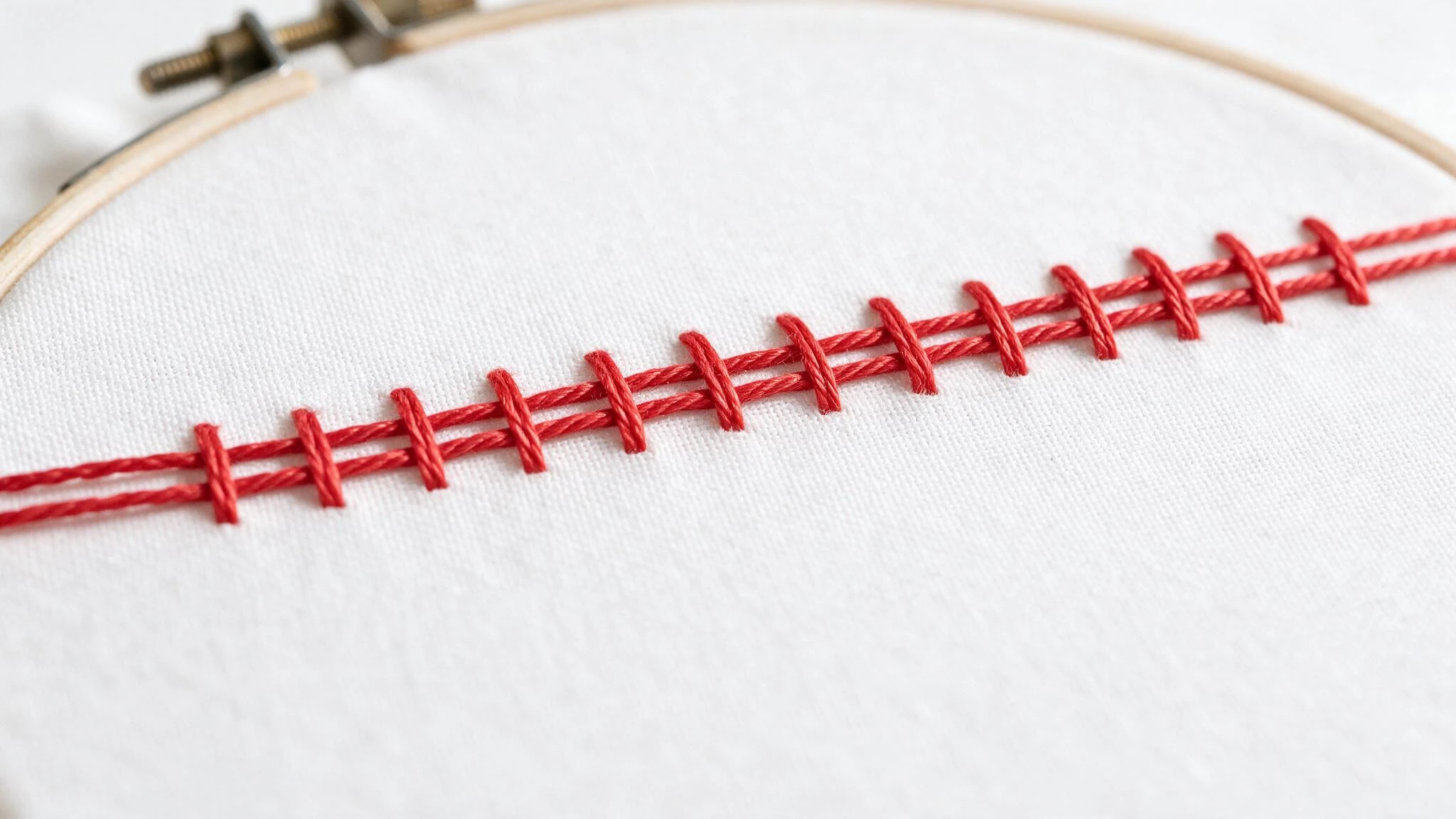 Couching stitch in red thread with parallel laid threads secured by evenly spaced short stitches across white fabric