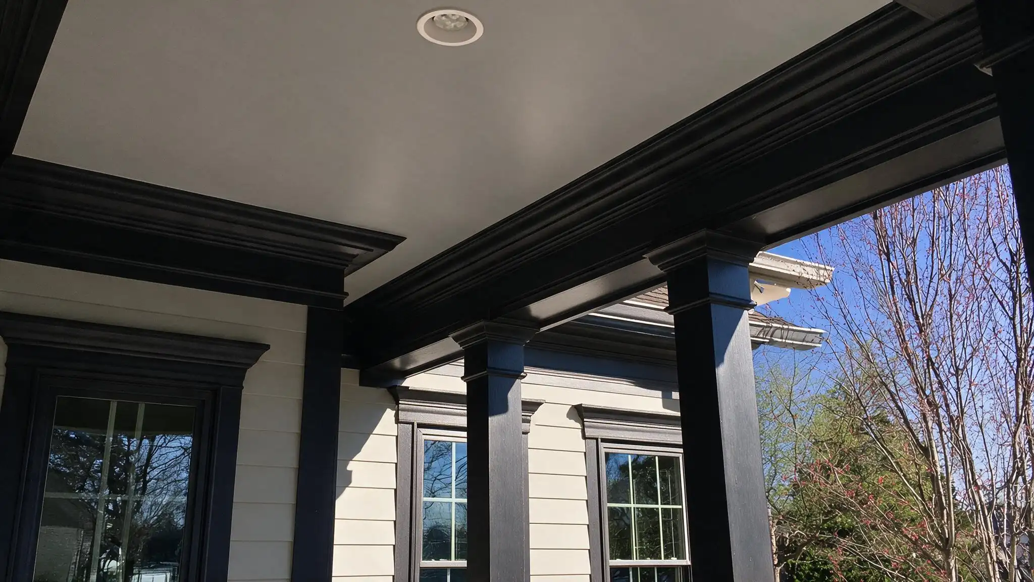 Covered porch with black columns and trim, cream siding, two windows, and a glass door beside leafless trees under a blue sky.