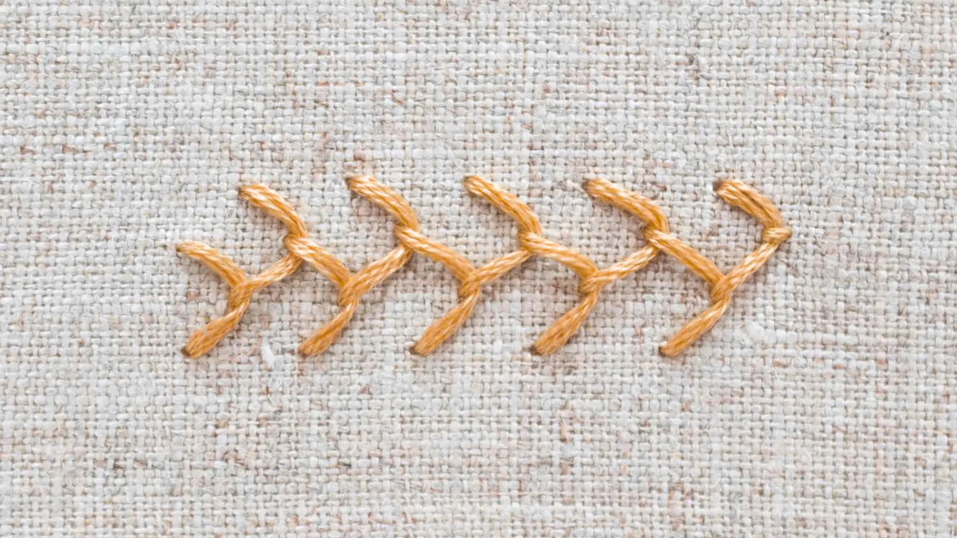 Feather stitch in orange thread forming a repeating branching decorative line across textured beige embroidery fabric