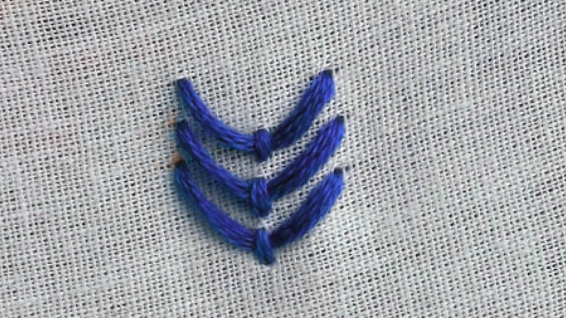 Fly stitch in blue thread forming three neat stacked V shapes with small anchoring stitches on textured beige fabric