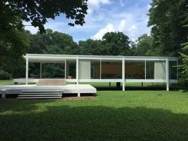 Front view of Farnsworth House Illinois featuring a glass walled steel framed modernist home elevated above a green lawn surrounded by tall trees