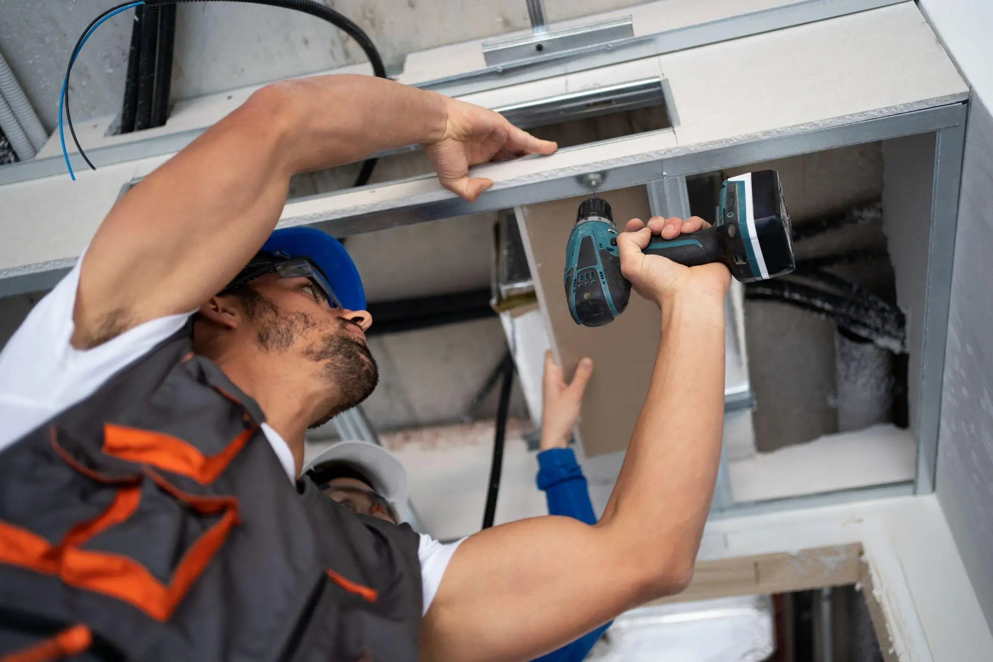 Worker in a blue hard hat and safety glasses using a drill on a ceiling frame