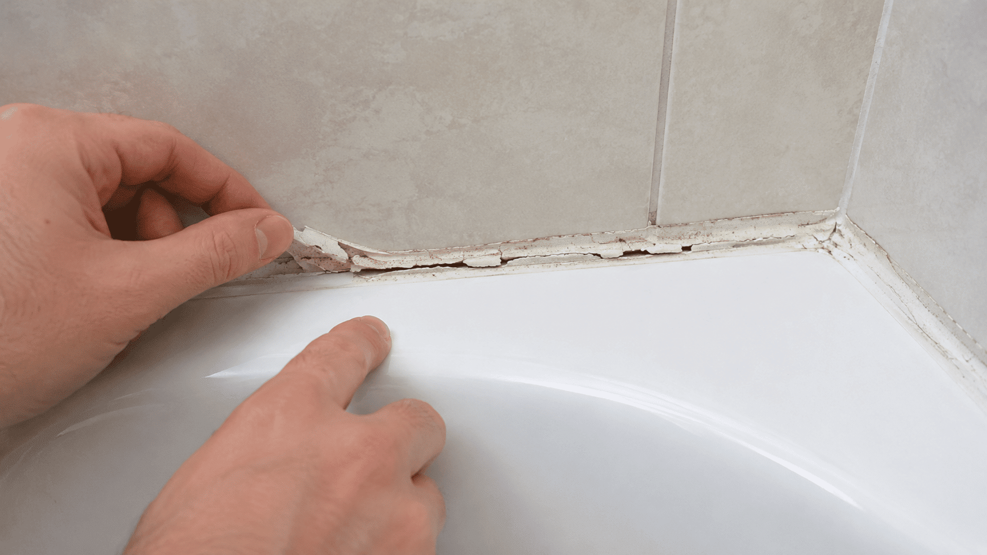 hands inspecting cracked and peeling caulk along the joint between a white bathtub and light wall tiles