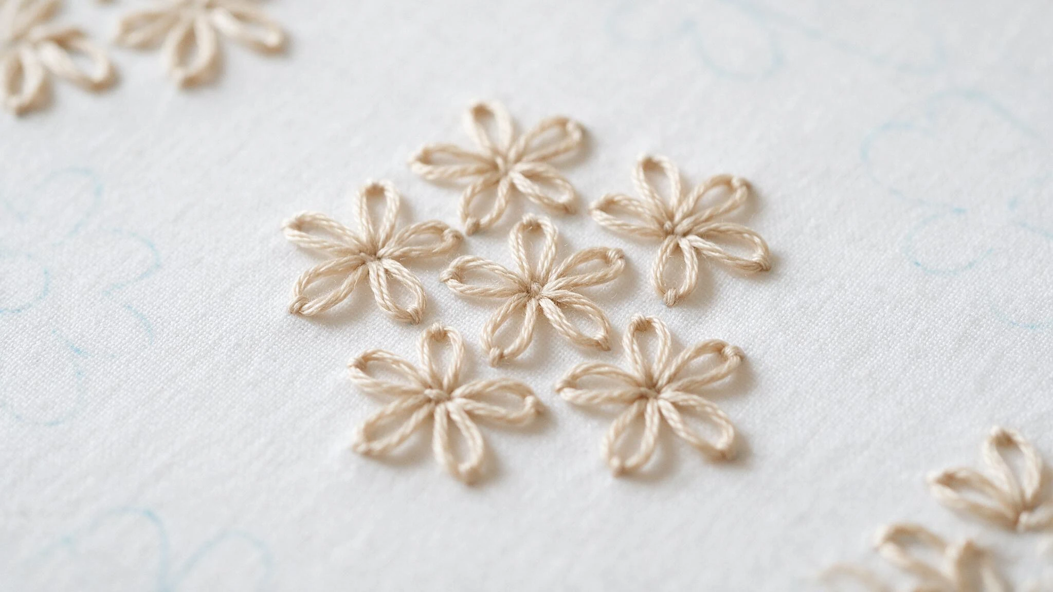 Lazy daisy stitch in beige thread forming small five petal flowers across white fabric in a simple embroidery sample