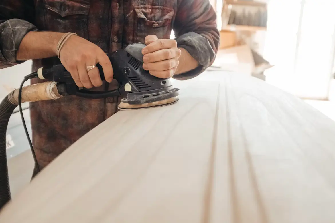 Person sanding wooden surface with power sander in a workshop wearing plaid shirt