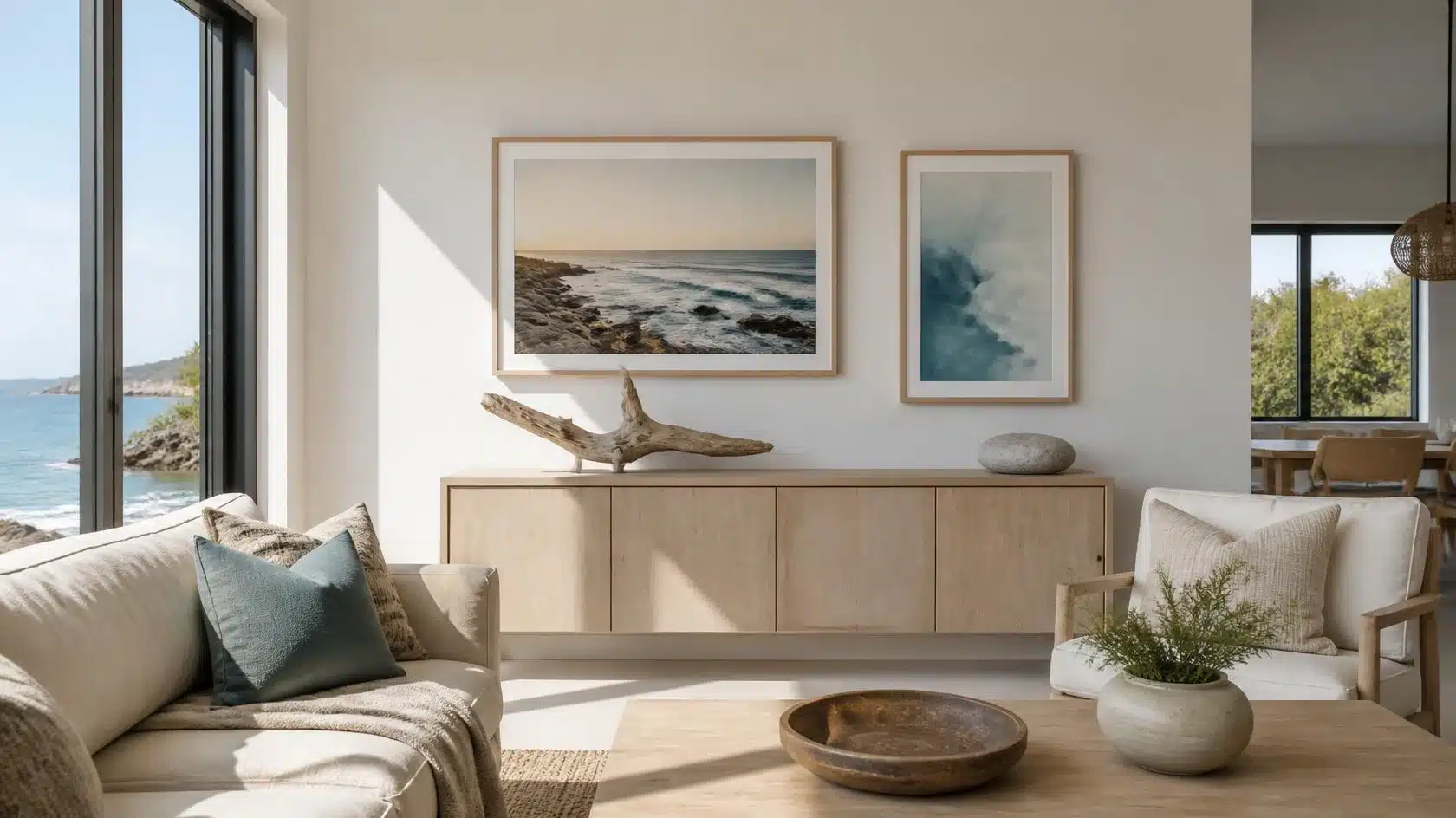 Modern beach house living room with framed coastal artwork, driftwood sculpture, oak sideboard, and ocean views through black-framed windows.