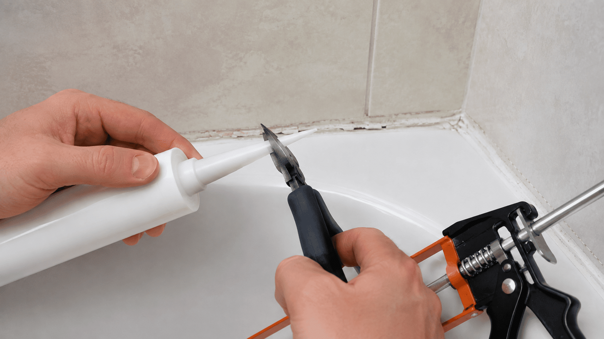 hands cutting the nozzle of a white caulk tube beside a caulk gun near a white bathtub and tiled wall