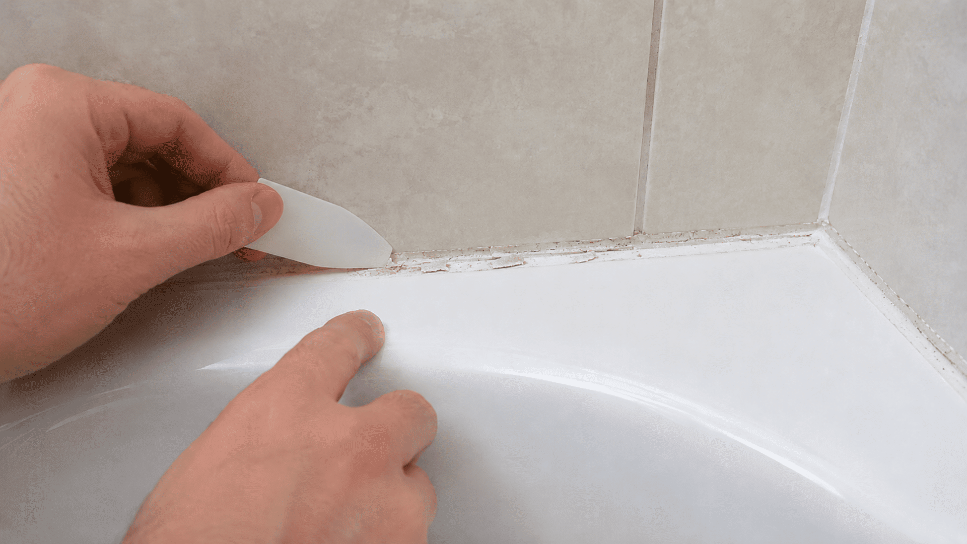 hands scraping leftover caulk residue from the joint where a white bathtub meets a light tiled wall