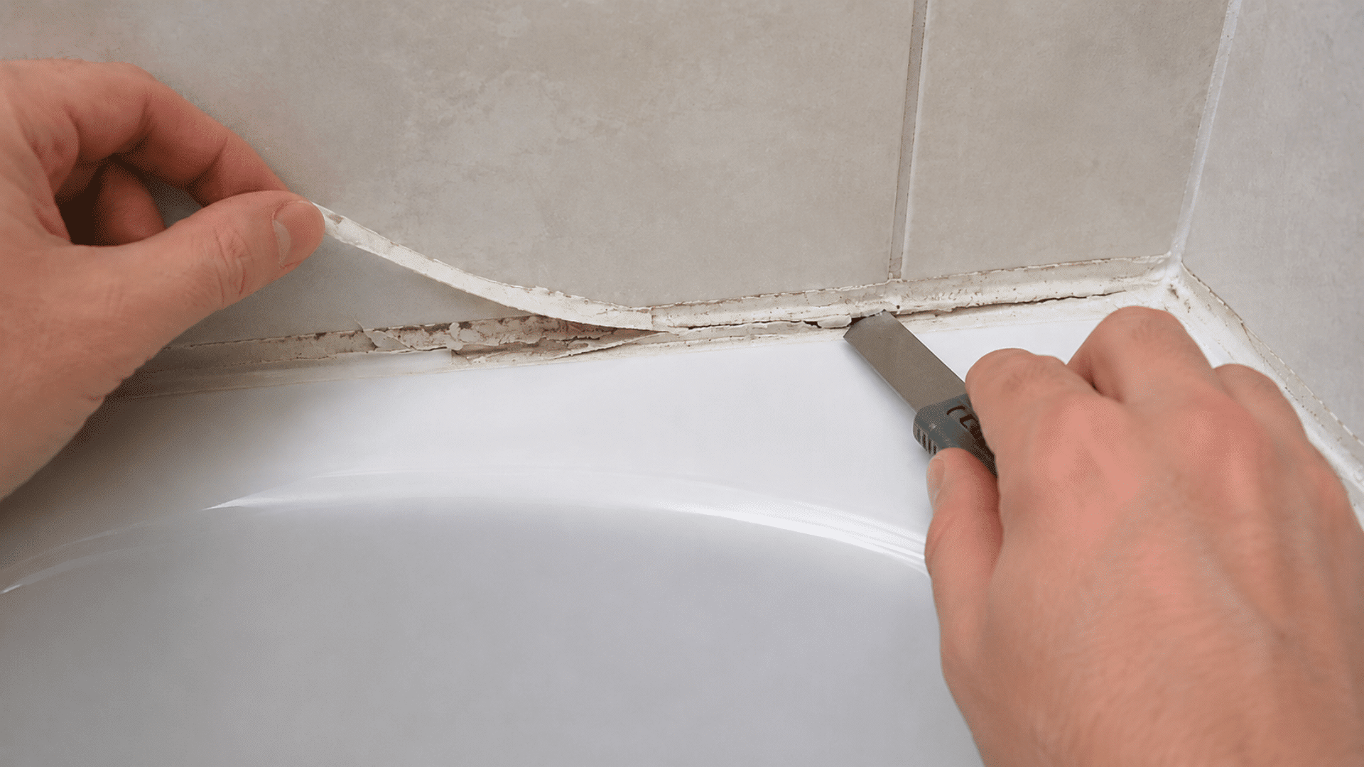 hands cutting and pulling old caulk from the seam between a white bathtub and light tiled bathroom wall