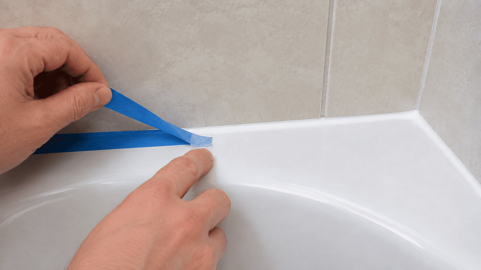 hands peeling blue painter's tape away from a smooth fresh white caulk line along a bathtub wall joint