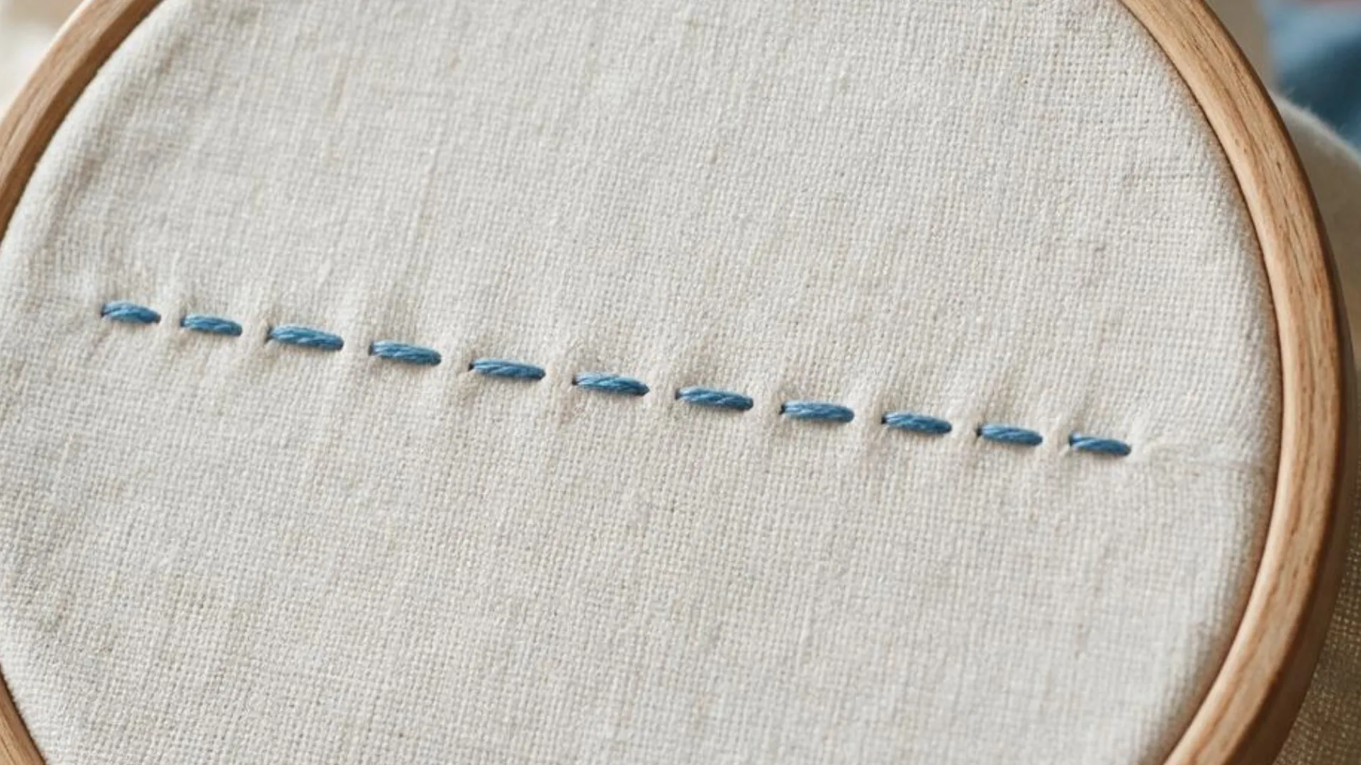 Running stitch in blue thread with evenly spaced short stitches across white fabric held inside a wooden embroidery hoop