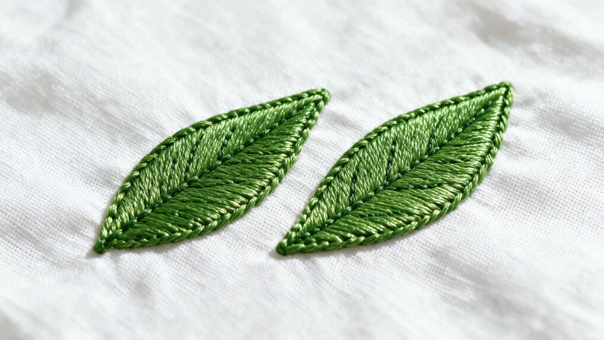Satin stitch in green thread filling two leaf shapes smoothly with dense glossy stitches across clean white fabric