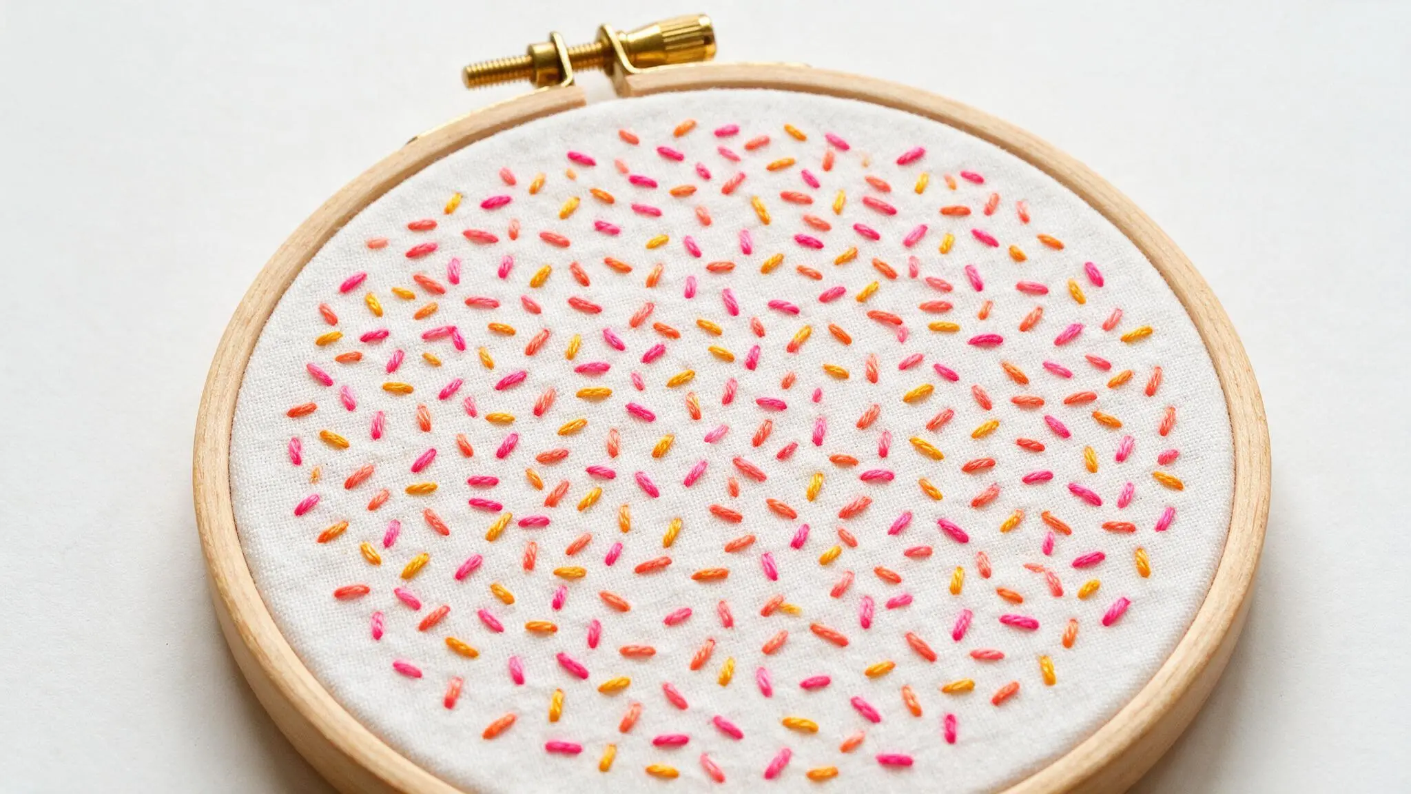 Seed stitch filling a wooden embroidery hoop with scattered short pink orange and yellow stitches on white fabric