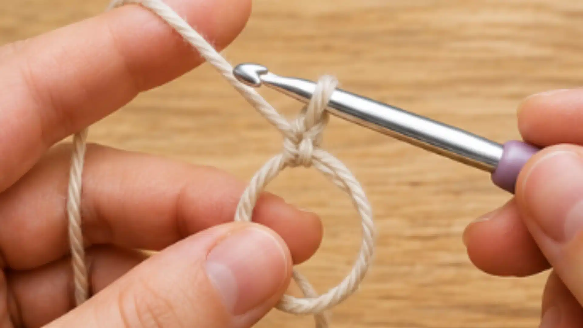 Close view of hands pulling beige yarn through a magic ring with a silver crochet hook over a smooth wooden table