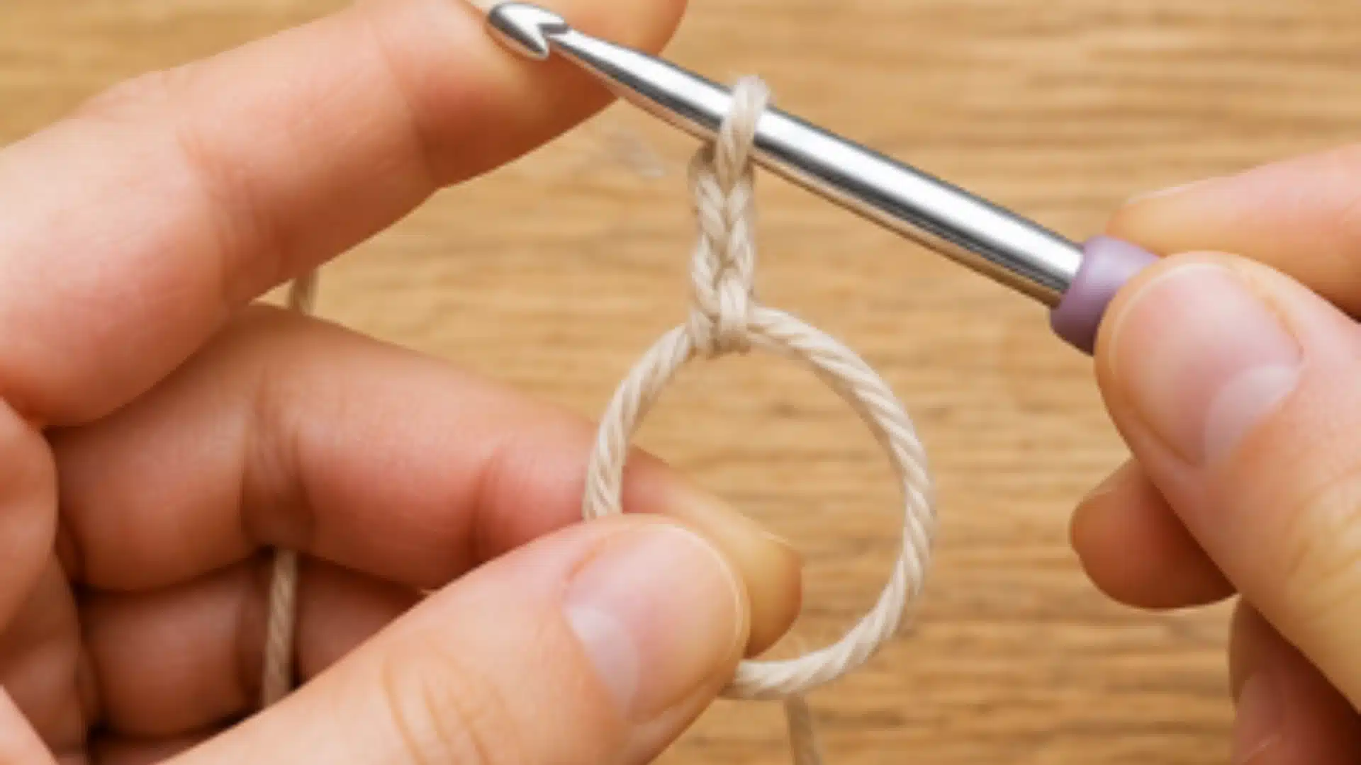 Hands making a chain above an open beige magic ring with a silver crochet hook on a light wooden surface indoors