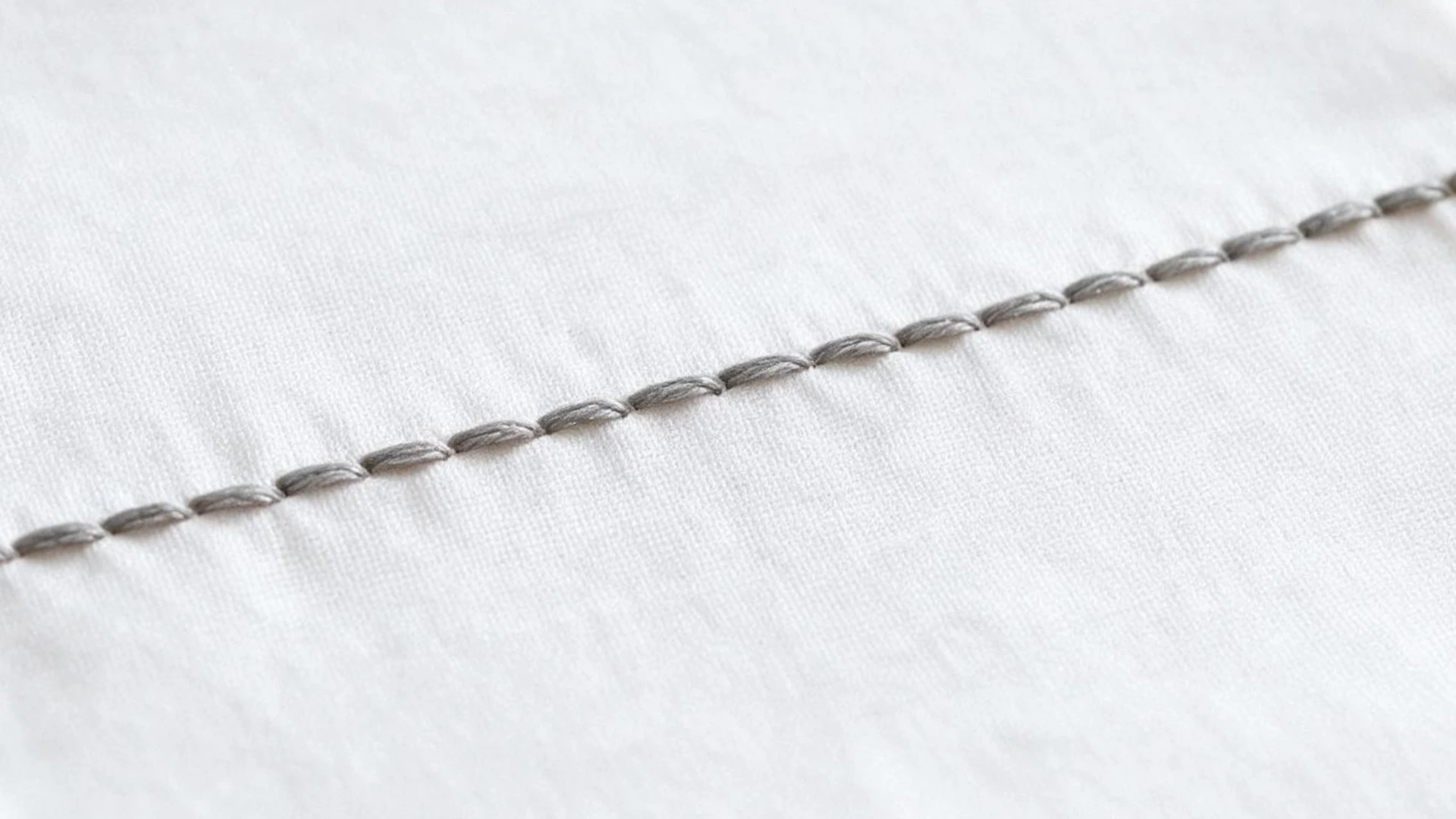 Straight stitch in gray thread forming a neat line of separate short stitches across smooth white embroidery fabric