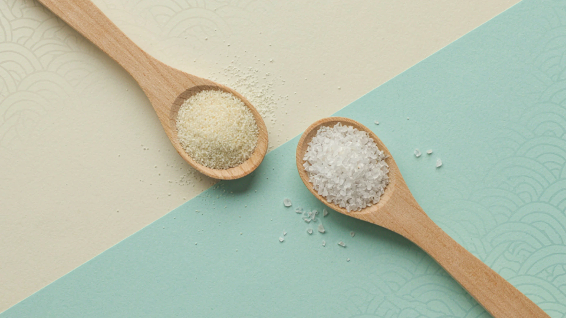 Two wooden spoons hold kosher salt and table salt on a diagonally divided beige and turquoise surface.