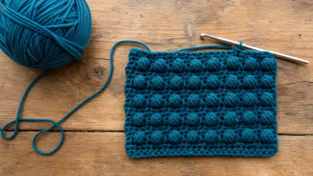 Deep teal bobble stitch crochet swatch with matching yarn ball and silver crochet hook on a rustic wooden table.