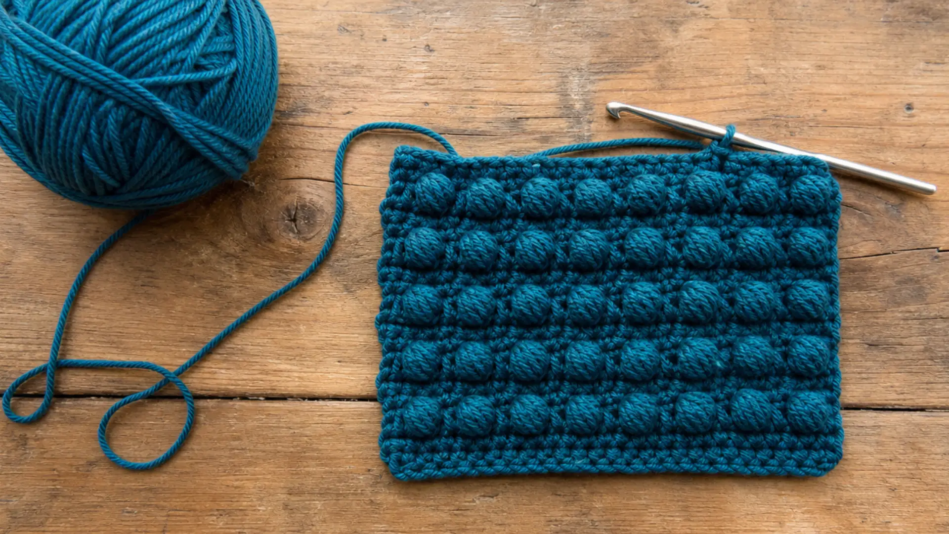 Deep teal bobble stitch crochet swatch with matching yarn ball and silver crochet hook on a rustic wooden table.