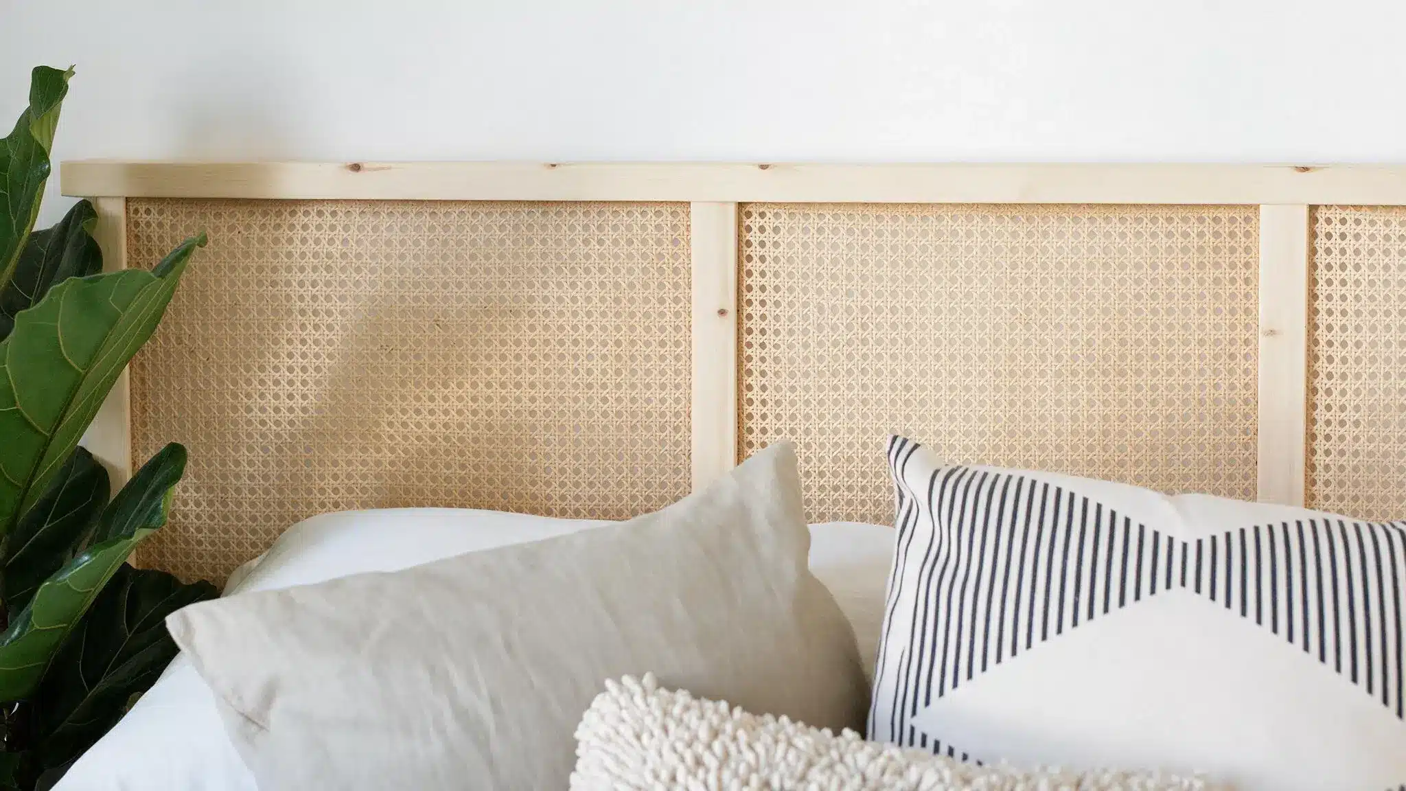 A bed with white and patterned pillows against a woven cane headboard, with leafy green plants along the left side.