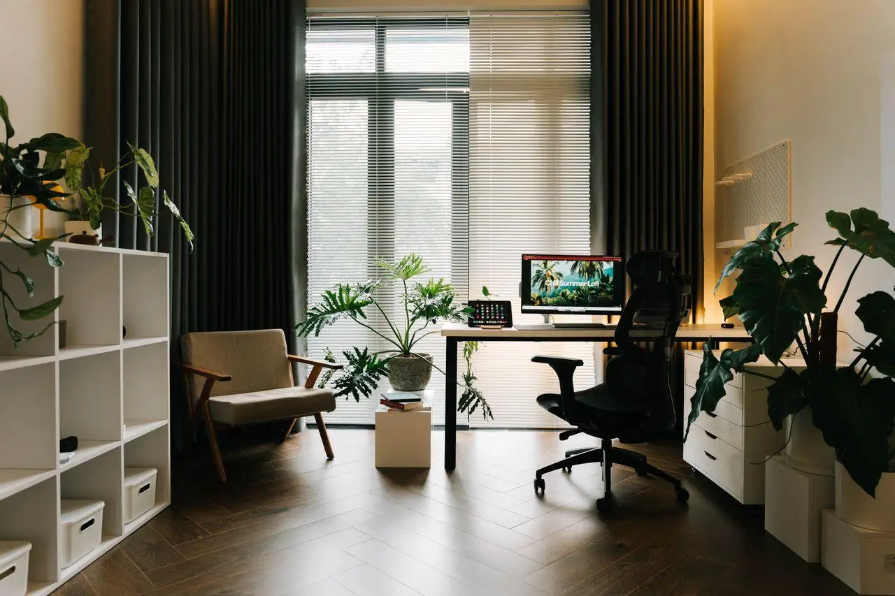 Modern home office with desk, computer setup, indoor plants, and armchair near large window blinds