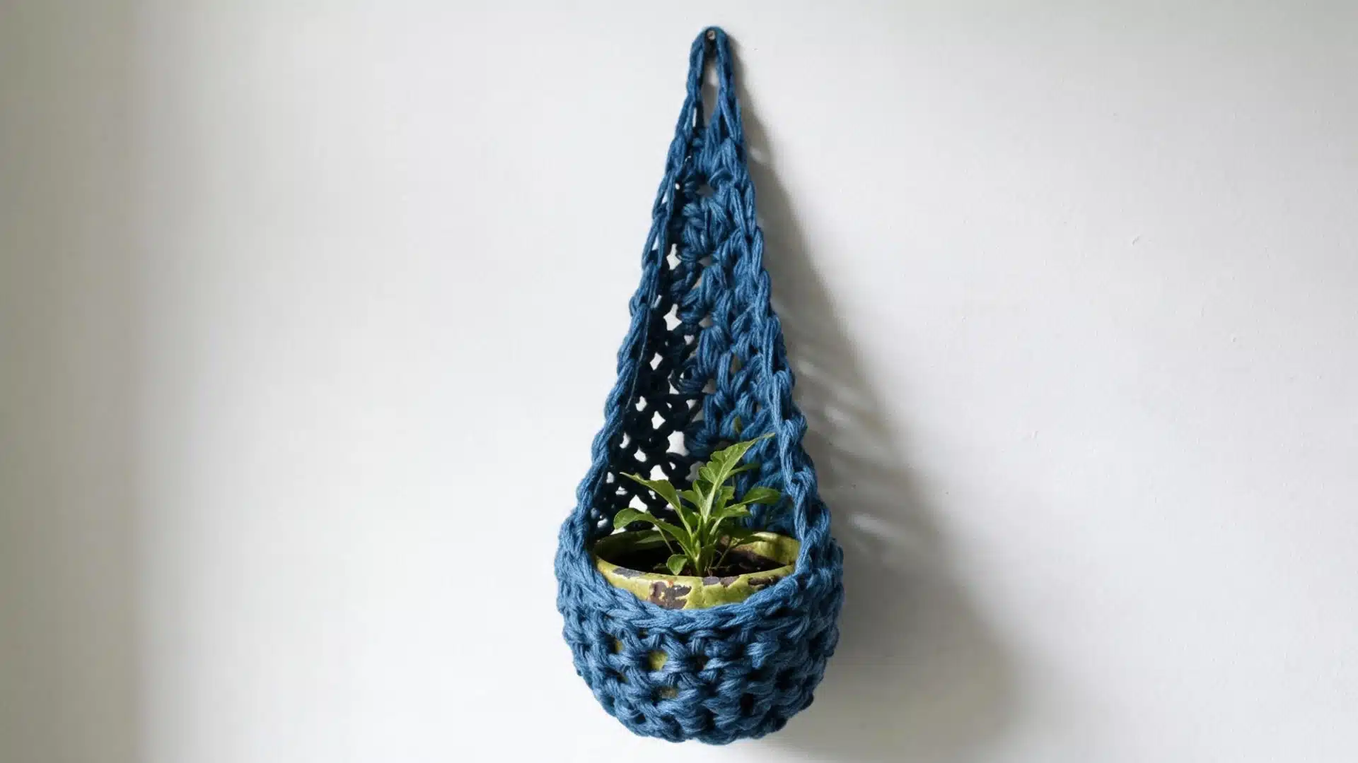 Blue crochet planter sack holding a small green plant in a patterned pot against a plain white wall.