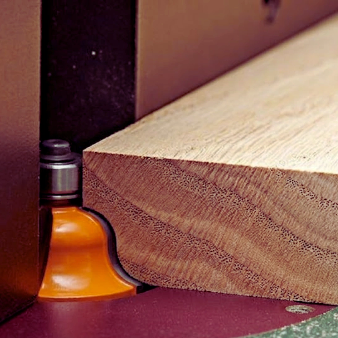 Close-up of a router table shaping a rounded edge on a thick wood board with an orange profile bit and bearing guide.