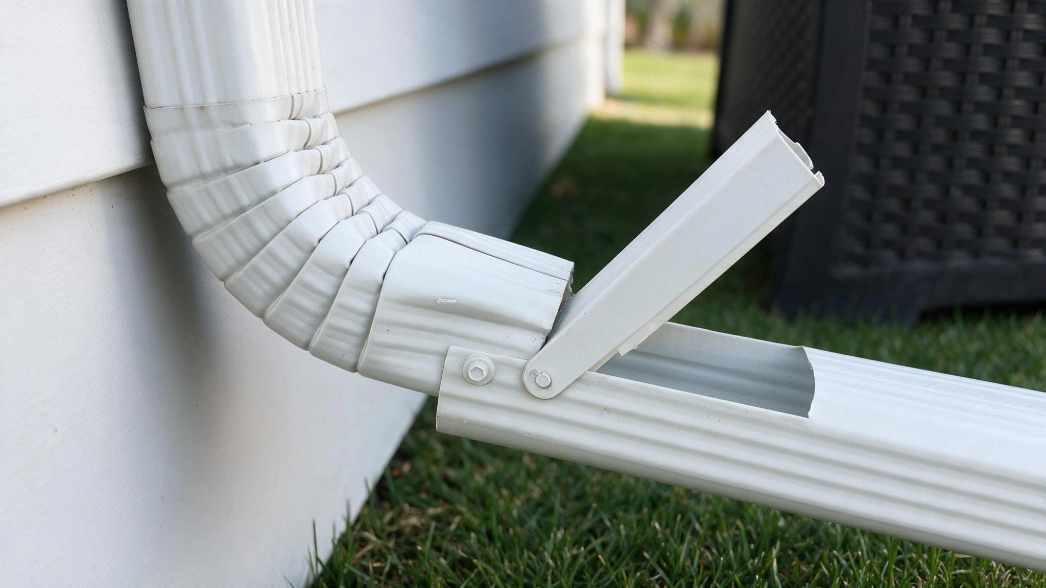 Close-up of a white vinyl gutter elbow and downspout section joined by a metal bracket beside a house wall and lawn.
